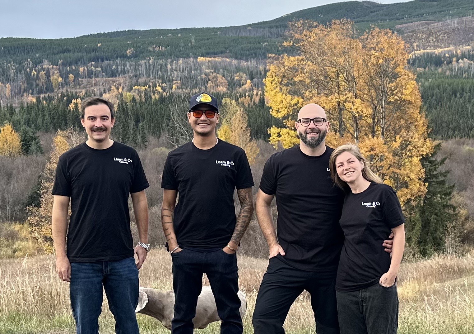 The four members of the Loam & Co. Catering team outdoors in their Loam shirts, with autumn trees and hills behind them